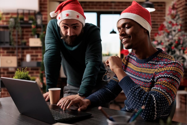 Men doing teamwork on laptop in decorated office, working on business in space with christmas tree. Celebrating seasonal tradition with festive decorations at company workplace.