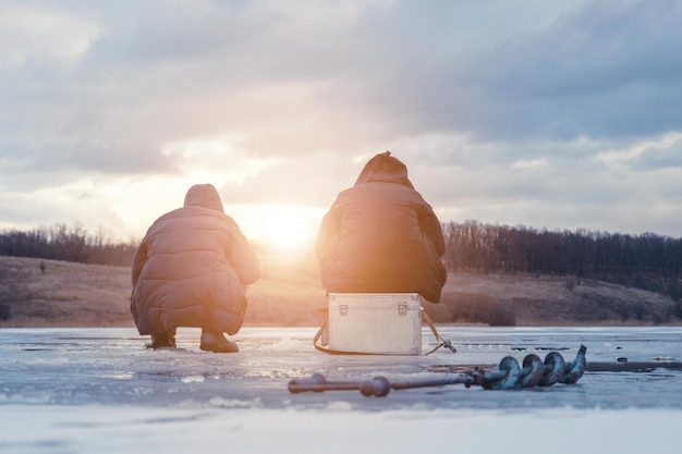 Men fishing on ice winter fishing on a frozen river in the evening at sunset