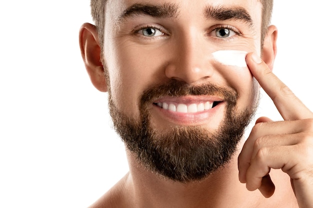 Men's beauty Young man is applying moisturizing and anti aging cream on his face against white background