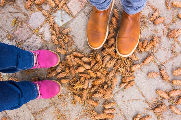 Men's brown boots and lilac women's boots on a path in the forest covered with cones of coniferous trees