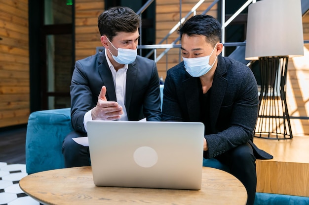 Men sit on the couch meet discussing joint business at a laptop computer wearing a face mask