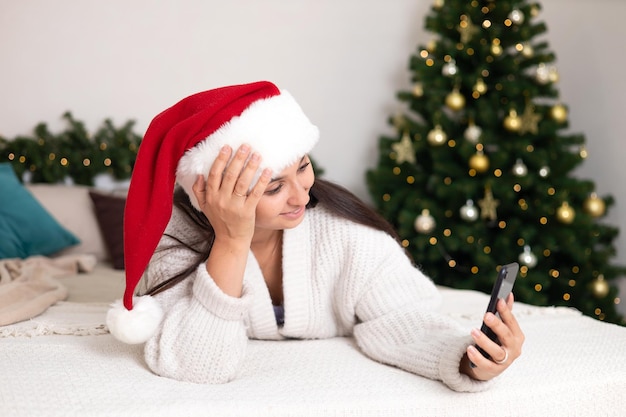 Merry Christmas A beautiful young woman in Santa39s hat makes a video call to her family lying in the bedroom with a decorated Christmas tree and lights in the background Lifestyle