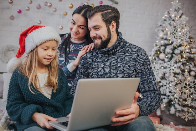 Merry Christmas and Happy Holidays Cheerful mom, dad and her cute daughter girl to use a laptop. Parent and little child having fun near Christmas tree indoors. Morning Xmas. 