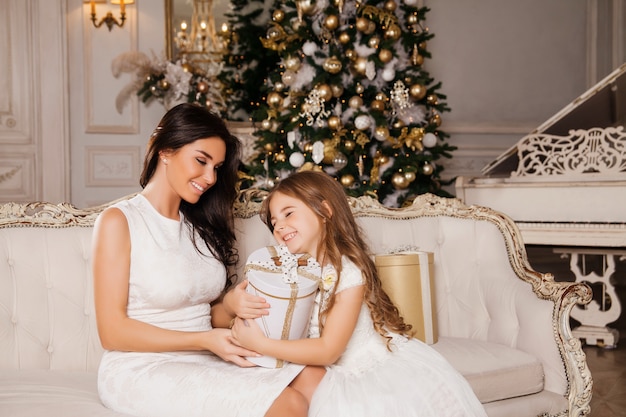 Merry Christmas and Happy Holidays. Cheerful mom and her cute daughter girl exchanging gifts in white classic interior  piano and a decorated Christmas tree. New Year 