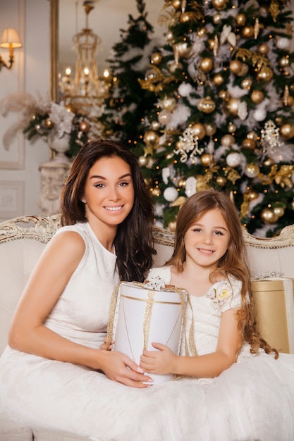 Merry Christmas and Happy Holidays. Cheerful mom and her cute daughter girl exchanging gifts in white classic interior  piano and a decorated Christmas tree. New Year 