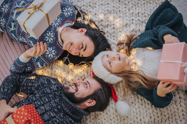 Merry Christmas and Happy Holidays Cheerful Mother, father and her cute daughter girl exchanging gifts. Parent and little child having fun near Christmas tree indoors. Morning Xmas. 