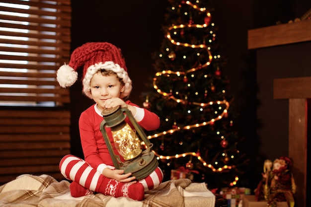 Merry Christmas and happy holidays A little boy is sitting with a lantern at the Christmas tree new year, elf.