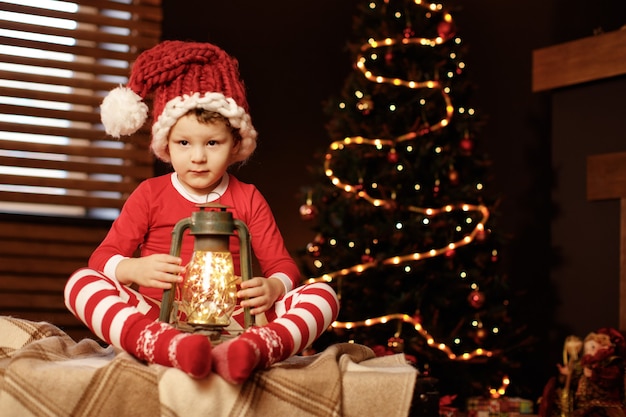 Merry Christmas and happy holidays A little boy is sitting with a lantern at the Christmas tree new year, elf.