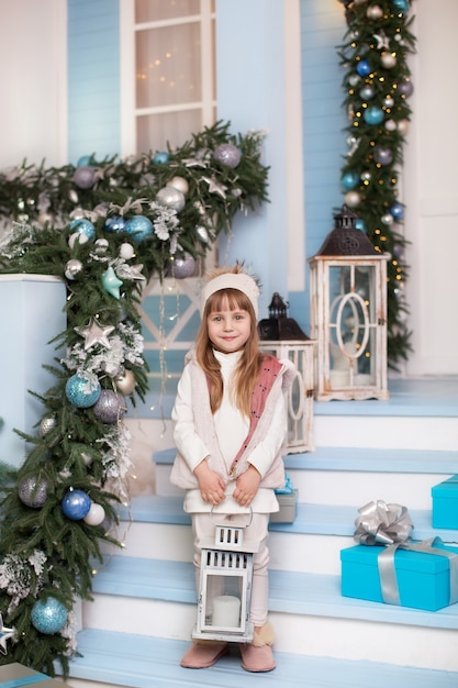 Merry Christmas, happy holidays! . little girl is standing with big Christmas lamp on porch of house. child decorates winter terrace for surface. beautiful child girl stands with lantern