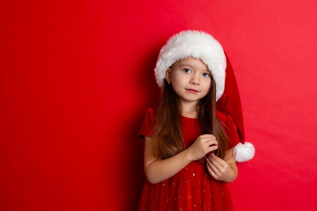 Merry Christmas and happy holidays. Portrait of a cute girl in a Santa cap on a red background. Kids, children. A place for text. High quality photo