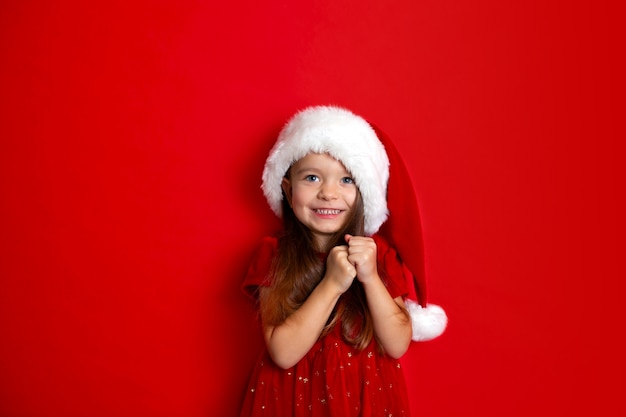 Merry Christmas and happy holidays. Portrait of a a pending happy girl in a Santa cap on a red background. Kids, children. A place for text. High quality photo