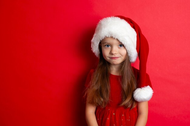 Merry Christmas and happy holidays. Portrait of a sad girl in a Santa cap on a red background. Kids, children. A place for text. High quality photo