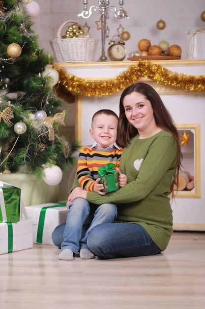 Merry Christmas and Happy Holidays. Pretty young mom with her fun little son near Christmas tree with gift boxes indoors.