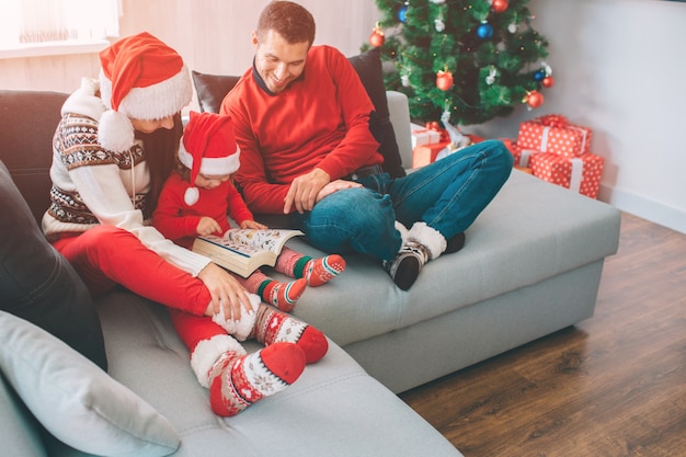 Merry Christmas and Happy New Year. Family sitting together on sofa. Small girl is in between her parents. She holds and looks at book with pictures. Man looks t it and smile. They are happy.