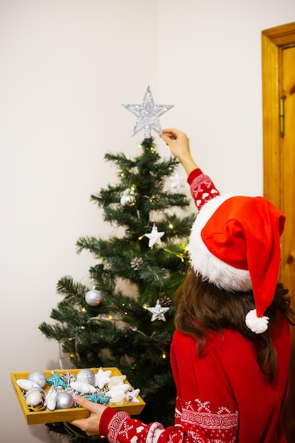 Merry Christmas and Happy New Year Female hands decorate the Christmas tree with balls and toys A woman in a New Year's hat all in red