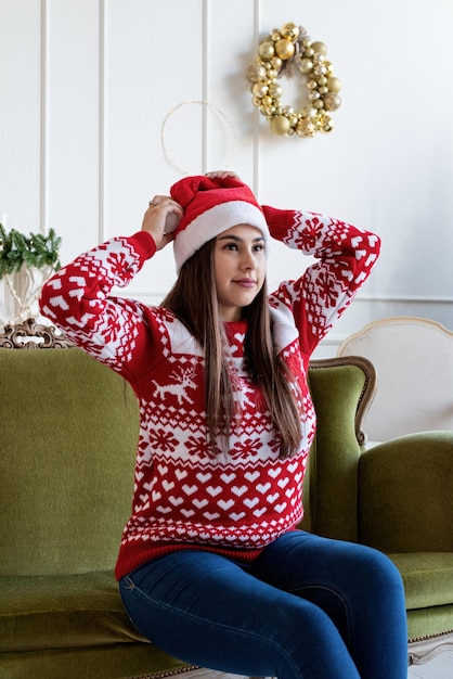 Merry Christmas and Happy New Year. Young brunette woman in santa hat sitting on green couch alone in a decorated for Christmas living room