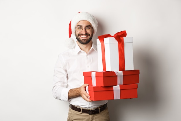 Merry christmas holidays concept happy young man smiling holding gifts in boxes and wearing santa ha...