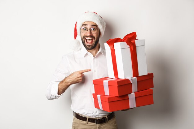 Merry christmas, holidays concept. Surprised man receive xmas gifts, pointing at presents and smiling happy, wearing santa hat, white background.