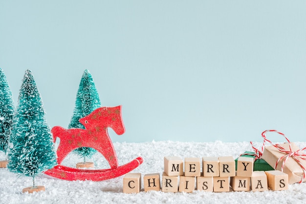 Merry christmas inscription with fir trees, horse toy, gift boxes on snow background