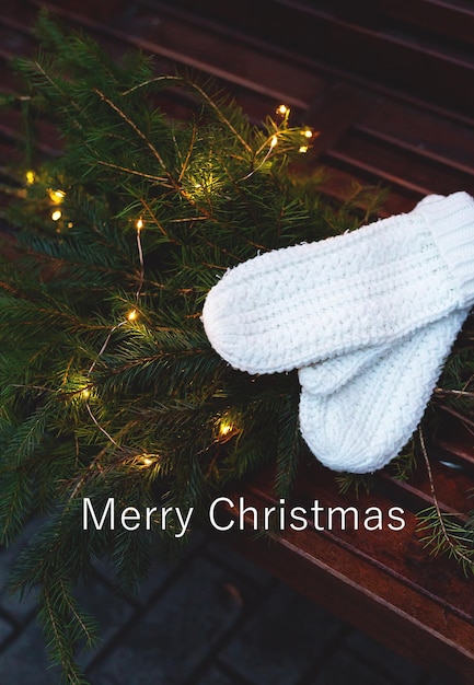 Merry christmas lettering Branches of a coniferous tree decorated with a garland lie on a bench along with knitted white mittens