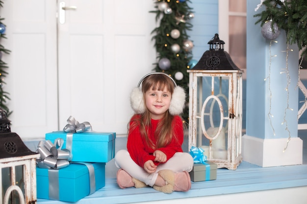 Merry Christmas! little girl is sitting in red sweater with gift on porch of house. child sits on a veranda decorated for Christmas. child opens a christmas present.