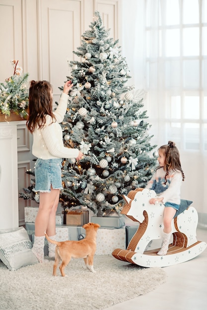 Merry Christmas! mom decorates the tree next to her daughter plays and runs a puppy