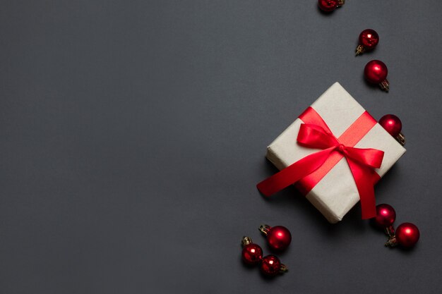 Merry Christmas and New Year greeting card with gift box wrapped in craft paper with red ribbon and balls on red background. Top view with copy space.