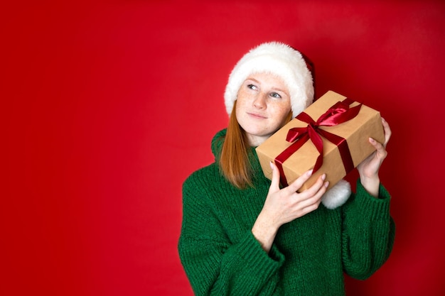 Merry Christmas Portrait of a beautiful young teenage girl in a cozy knitted green sweater and Santa39s hat holding gift boxes The red background is the place for the text