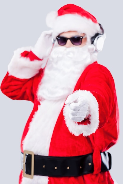 Merry Christmas to you! Cool Santa Claus in sunglasses adjusting his headphones and pointing you while standing against grey background