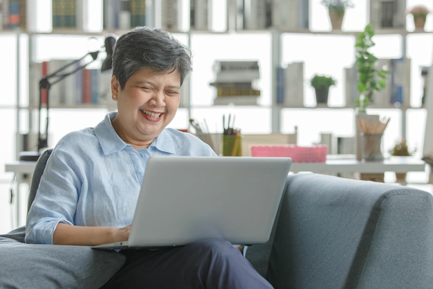 Merry middle aged Asian female with short hair smiling and browsing netbook while resting on couch in modern sunlit living room