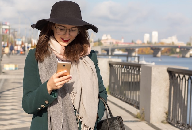 Merry smiling brunette woman in glasses and hat dressed in fashionable clothes using mobile phone