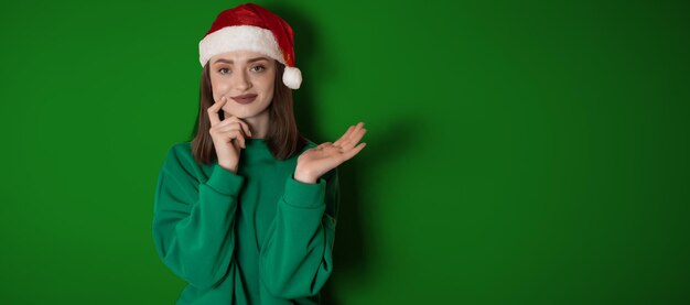 Merry surprised young woman wear xmas sweater Santa hat posing pointing hands arms aside indicate on workspace area isolated on plain pastel light green background Happy New Year 2023 holiday concept