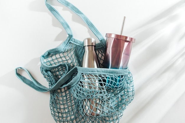 Mesh bag with reusable metal water bottle and cup on white background.   