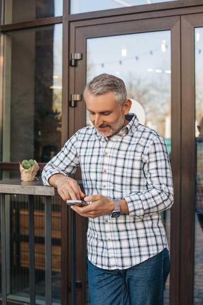 Message. Gray-haired middle aged man smiling looking into smartphone standing waiting outdoors near building during daytime