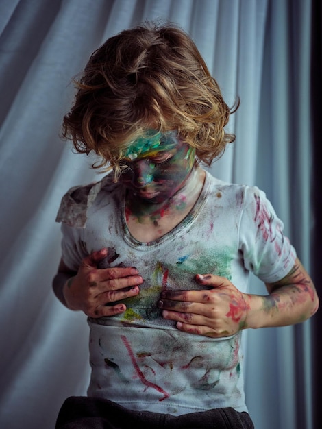 Messy boy with curly hair and painted face wiping dirty hands on messy t shirt while standing in light room