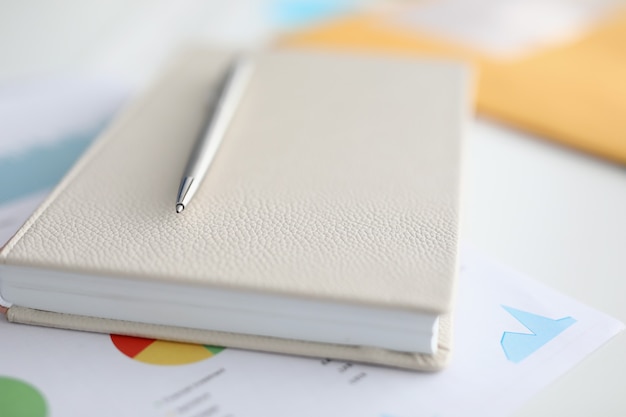 Metal ballpoint pen lying on notebook and documents with charts and diagrams closeup