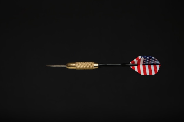 Metal dart for playing darts with color plumage under US flag, on black background.