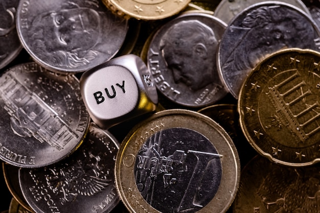 A metal dice in a pile of coins from different countries. On the edge of the dice is the word "buy". The concept of buy-sell choice.