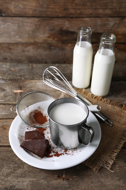 Metal mug and glass bottles of milk with chocolate chunks and strainer of cocoa on plate with burlap cloth and rustic wooden planks background