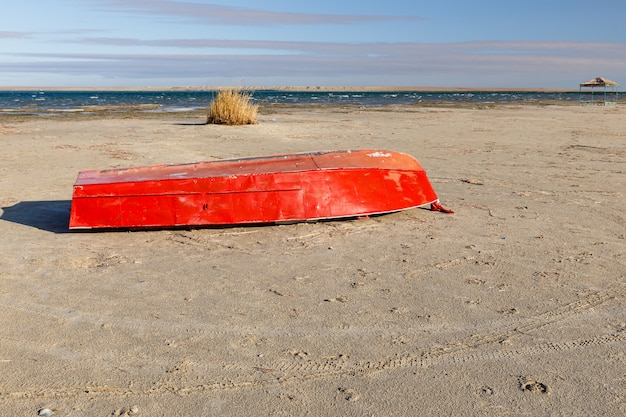 Metal red boat lies on the sand by the lake