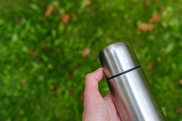 A metal thermos with a mug lid lies in the hand in the garden against the background of a lawn with green grass and rarely scattered dry leaves.