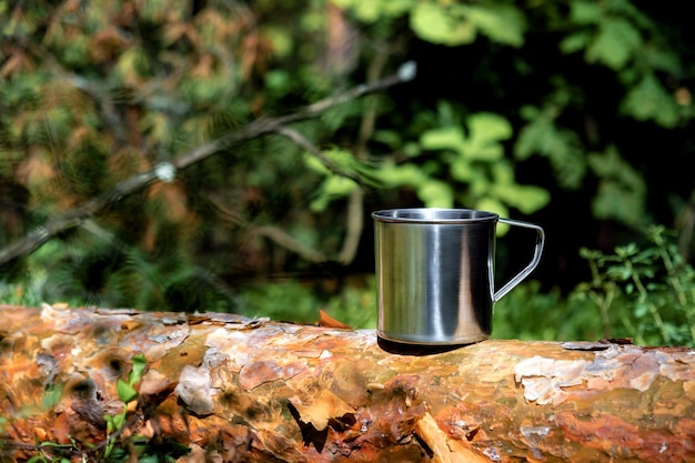 Metal tourist mug with hot drink stands on tree trunk in the forest