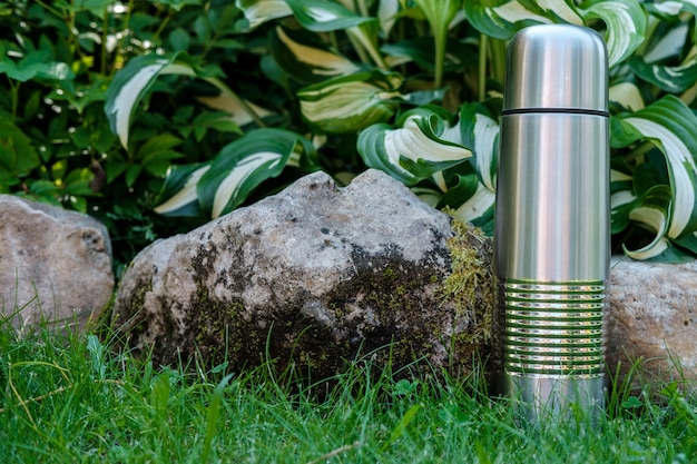 A metal tourist thermos for drinks stands on the grass among the stones covered with moss and large leaves in the background.