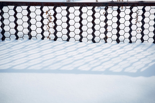 Metallic fence casts a shadow on the pure white snow. Calmness in the park after snowfall. Abstract winter background.