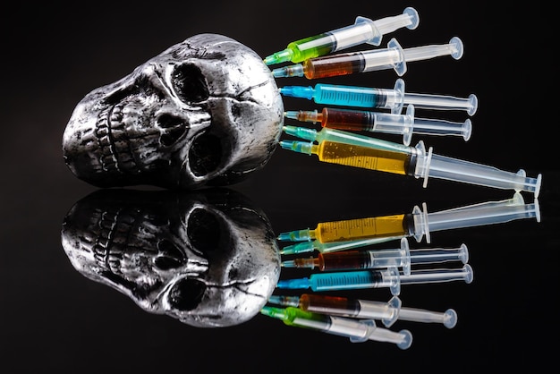 Metallic humans skull with colorful stucked syringes on black background reflective mirror surface d