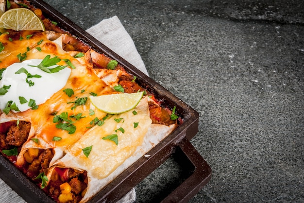 Mexican food. Cuisine of South America. Traditional dish of spicy beef enchiladas with corn, beans, tomato. On a baking tray, on a black stone . 