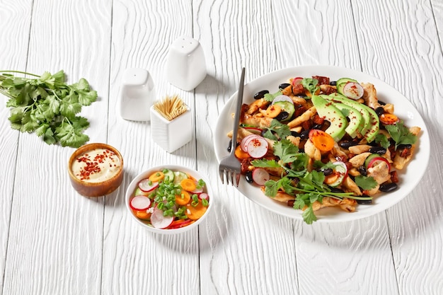 Mexican salad of black beans chicken cilantro radish carrot cucumber and avocado with tahini dressing and chili flakes served on white plate on white wooden board with cutlery top view closeup