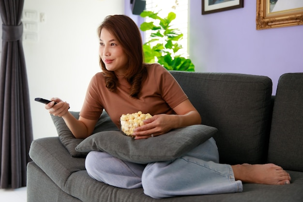 Mid adult Asian woman sitting on sofa in home living room holding bolw of popcorn and using remote control aiming to tv while watching in relax time.