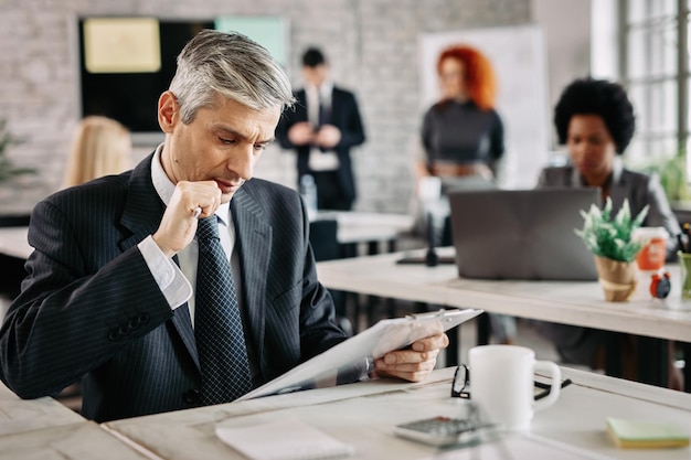 Mid adult businessman sitting in the office and reading data from clipboard