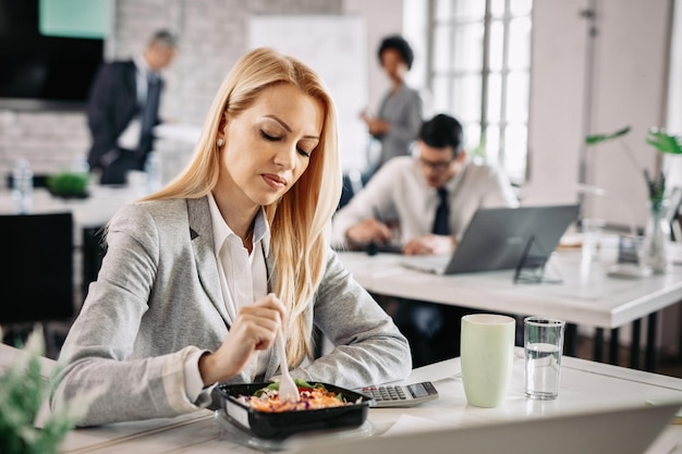 Mid adult businesswoman having lunch break at work and eating vegetable salad at her desk